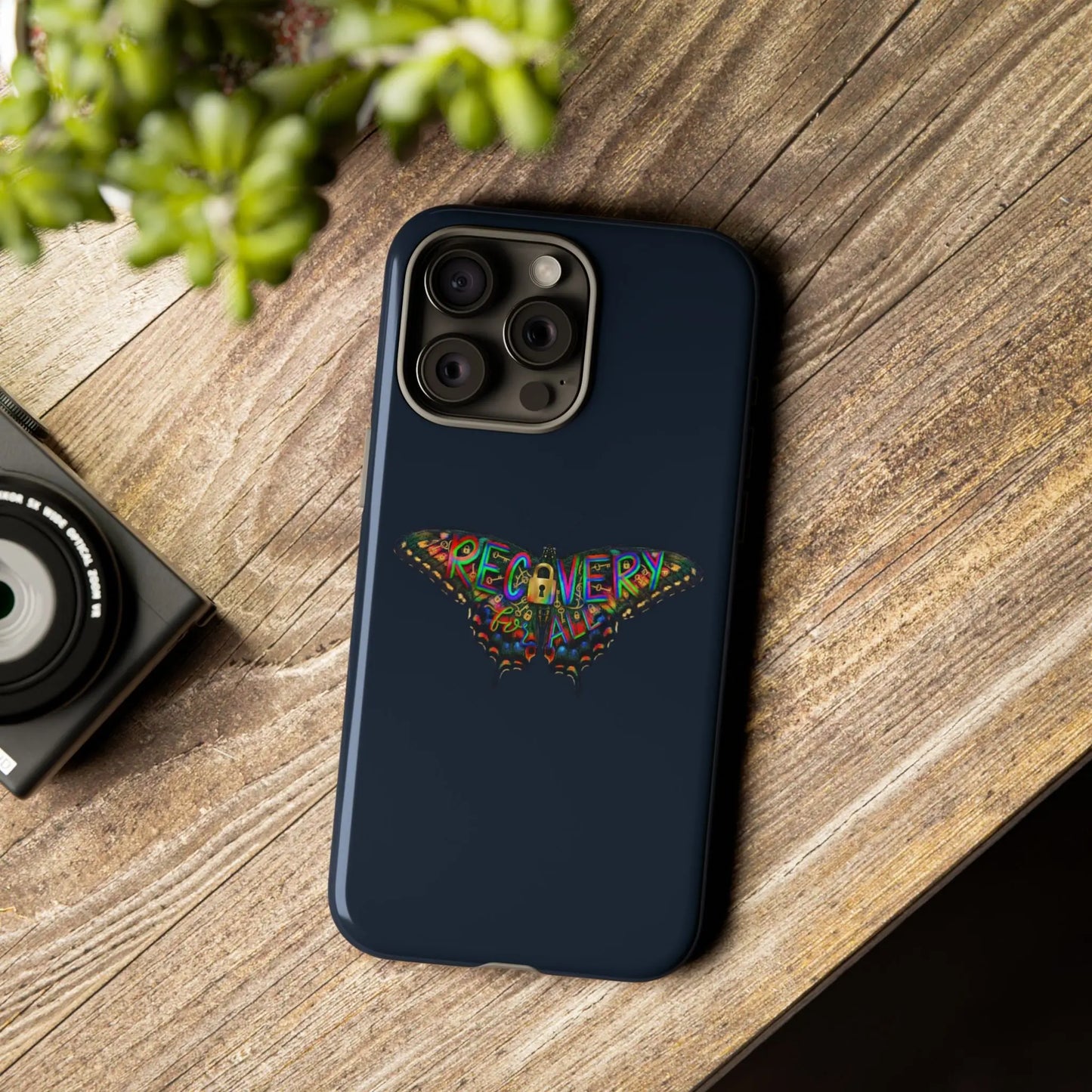 Navy blue phone case with a Recovery Butterfly design on a wooden surface next to a camera and plant.